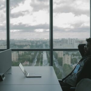 A man sits in an office with hands on head in front of a laptop, overlooking a cityscape.